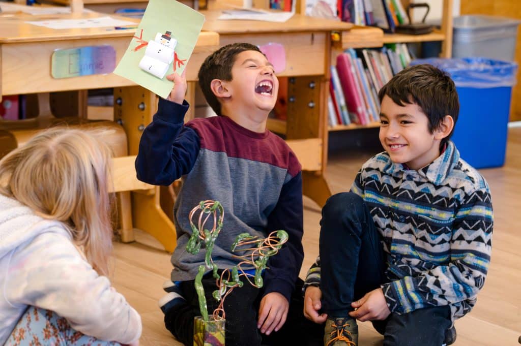 2nd or 3rd grade boy laughing while holding a card sitting next to two other students