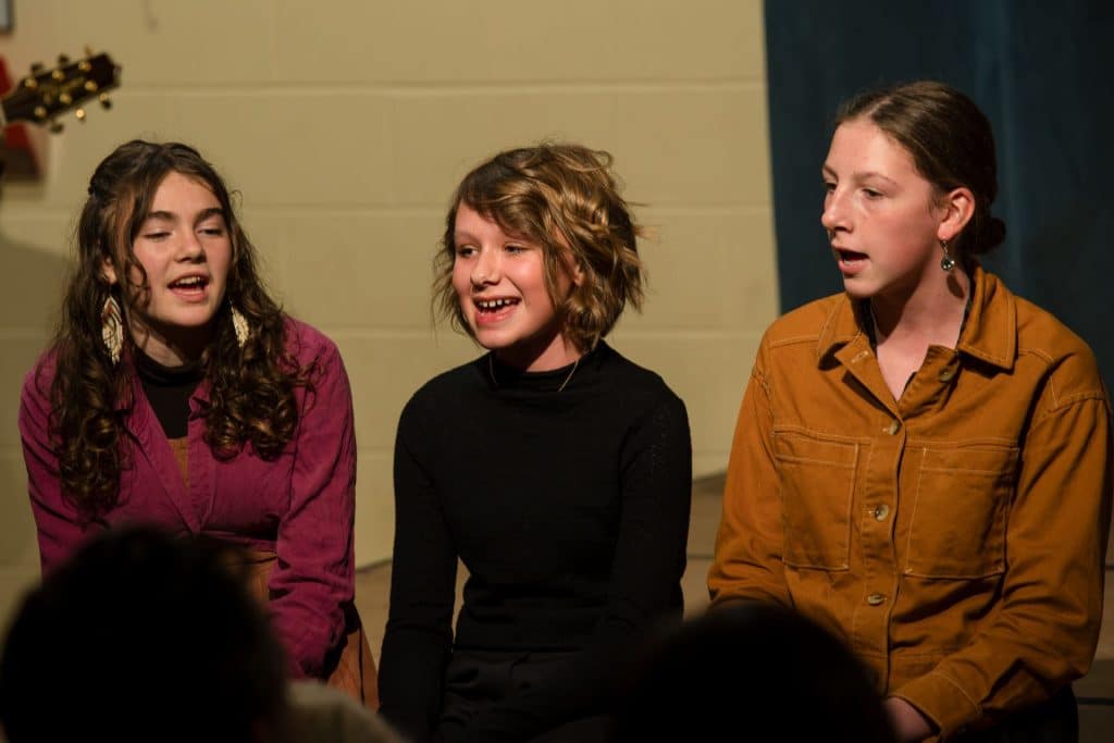 three middle school girls sitting on the edge of a stage and singing