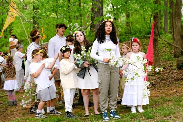 group of mixed age students wearing white clothes, holding flowers walking through a forest together